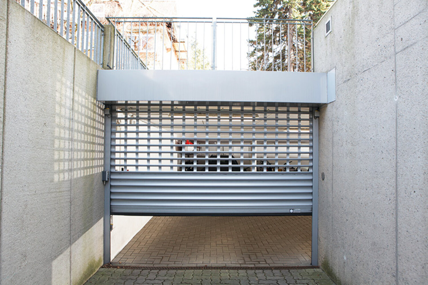 Ansicht eines halb heruntergefahrenen, alufarbenden Rolltores an einer Garageneinfahrt mit seitlichen Sichtbetonwänden