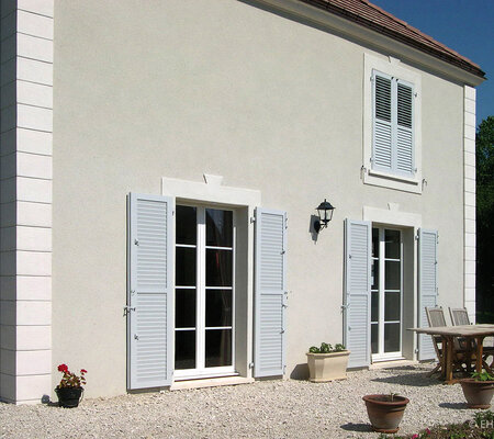 Schutz und Akzent: Haus mit bodentiefen Fenstern mit klassischen weißen Klappläden, beim Fenster im 1. O.G. geschlossen.